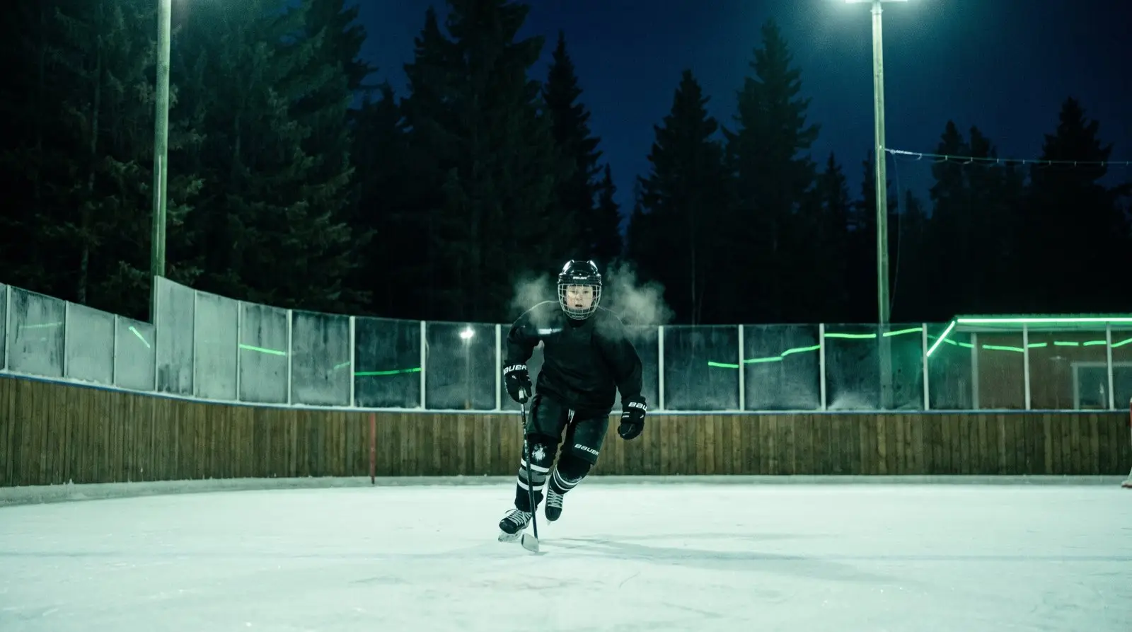 A young player in full gear skating up the open-air rink at night